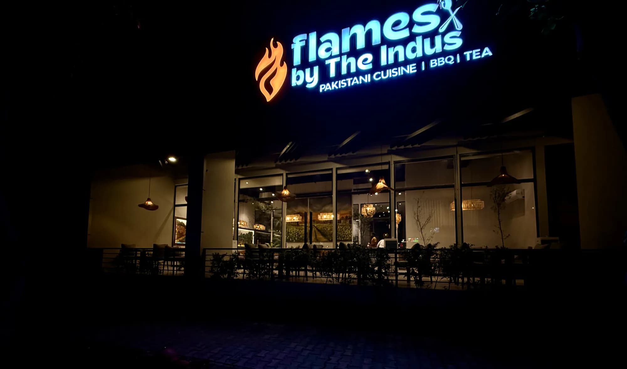 The lit Flames by the Indus sign above the restaurant's glass frontage at night
